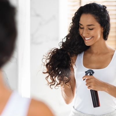 Smiling woman with curly hair, applying a protective hair formulation from a spray bottle to her ends.