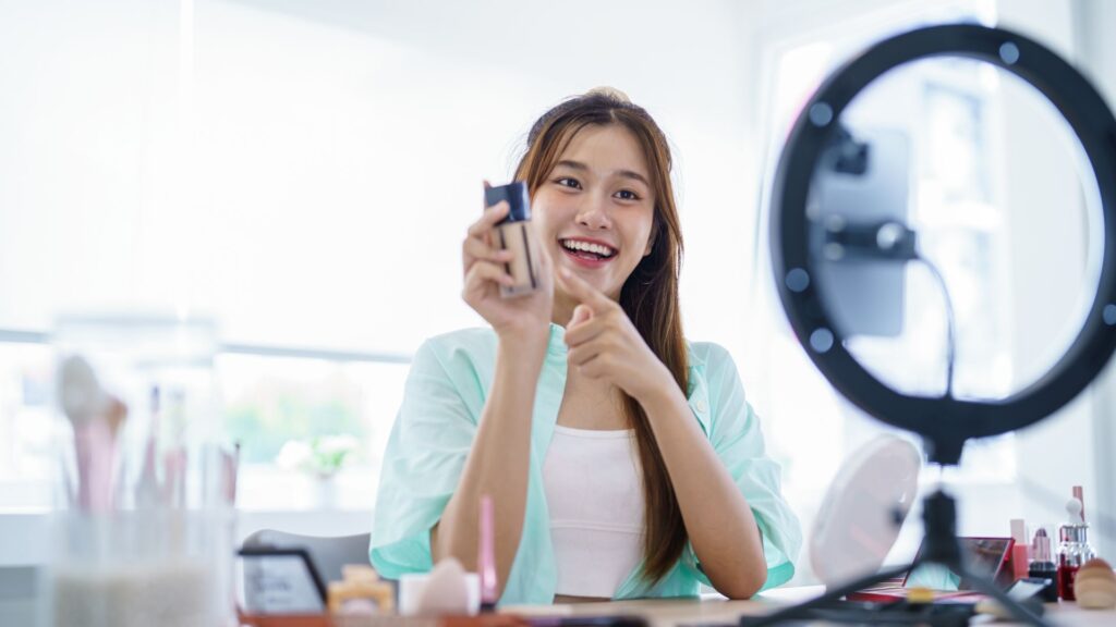 Woman sitting at a table with beauty products, using a smartphone on a tripod with a ring light, depicting AI beauty technology being used to explore products at home.